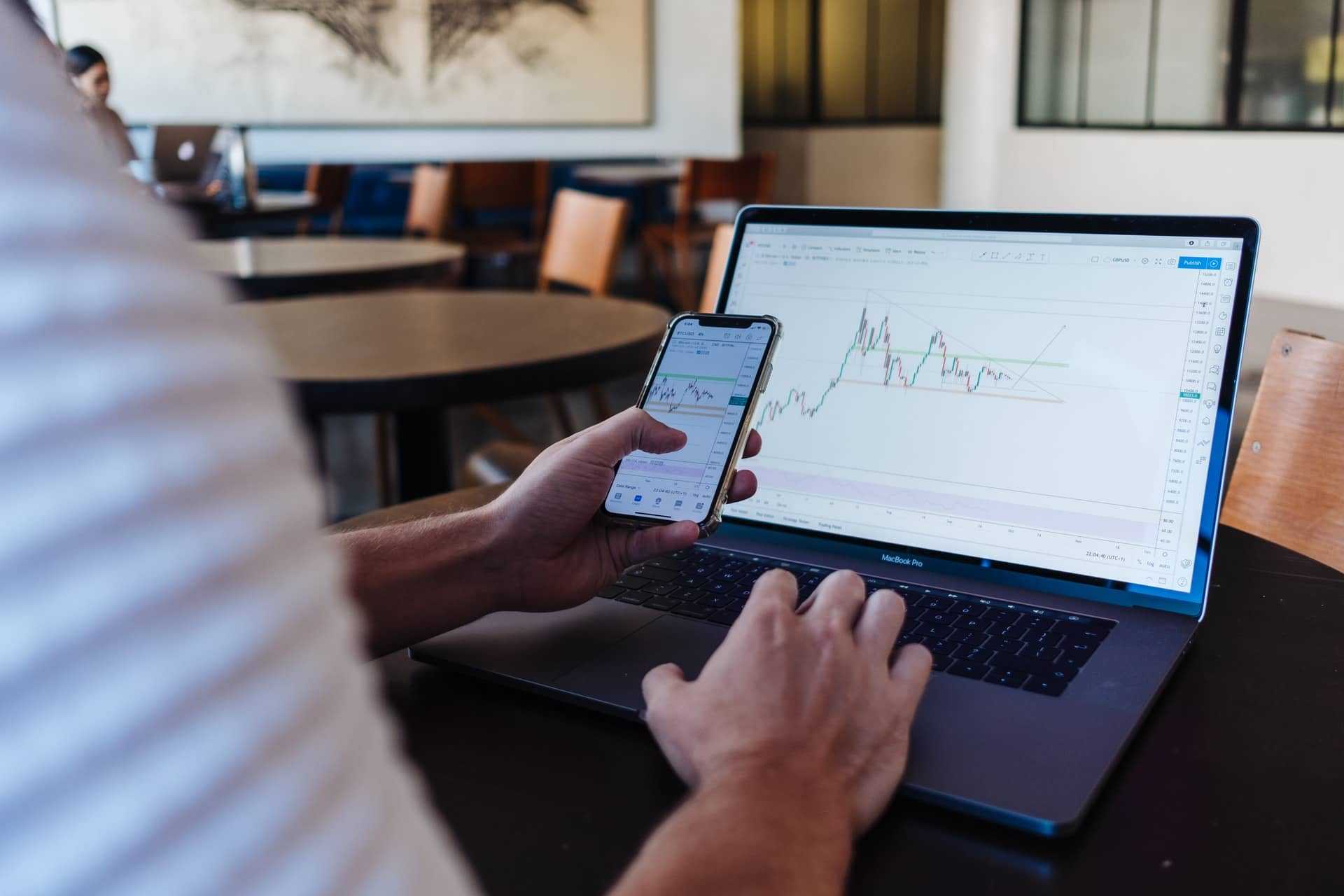A man sitting at a desk looking at a stock trading chart on his phone and laptop
