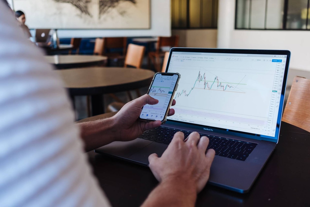 A man sitting at a desk looking at a stock trading chart on his phone and laptop
